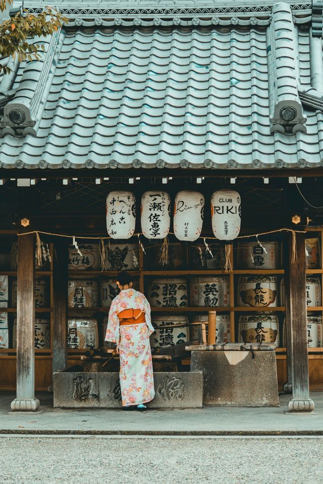 A woman in a floral kimono, seen from behind, stands at a water basin in front of a Japanese shrine with hanging lanterns.