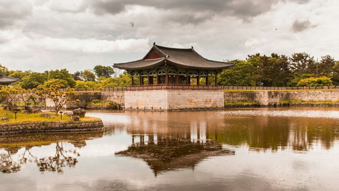 A traditional Korean pavilion on a stone foundation sits over a calm lake, its reflection visible in the water under a cloudy sky.