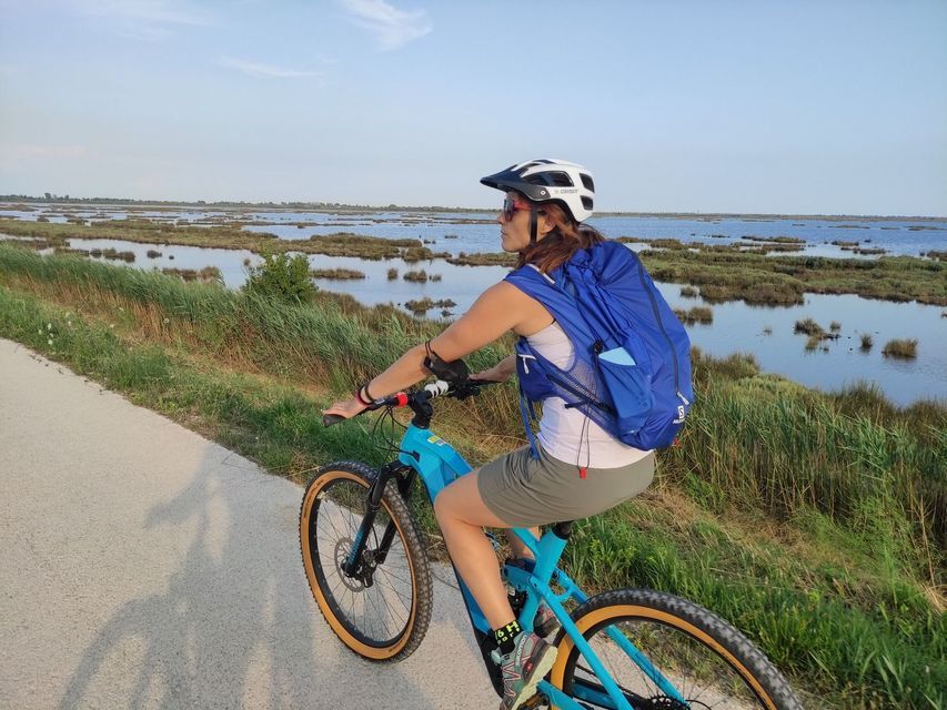 Una donna con casco e zaino percorre un sentiero in bicicletta blu acceso, accanto a una grande palude, sotto un cielo sereno.