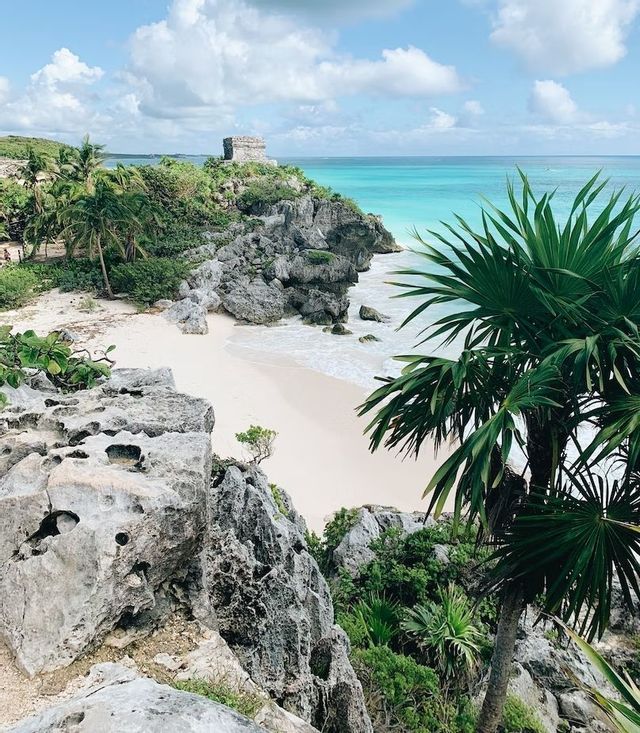 An ancient stone ruin on a rocky cliff overlooks a white sand beach and a turquoise sea, with palm fronds in the foreground.