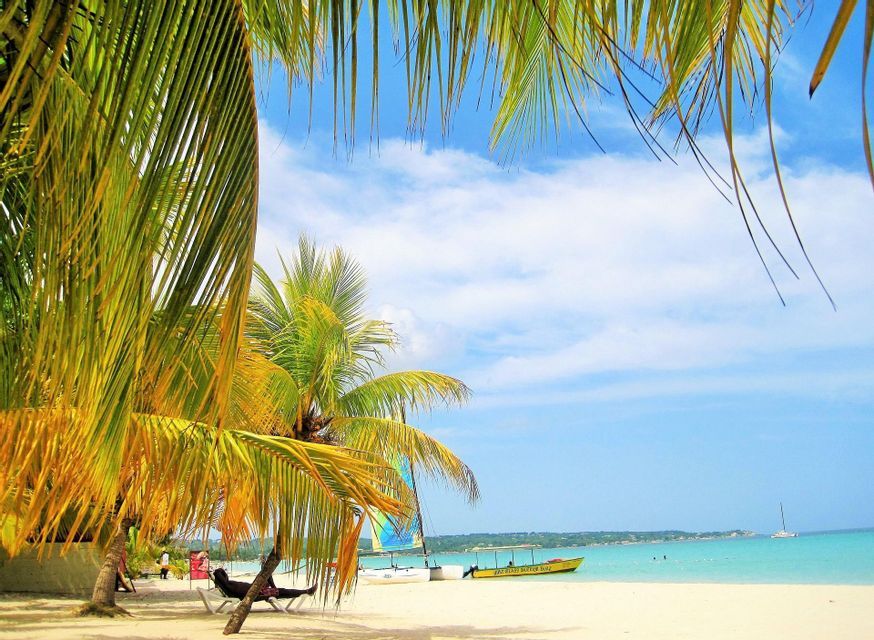 Fronde di palma incorniciano una spiaggia di sabbia bianca con barche su acqua turchese sotto un cielo parzialmente nuvoloso.