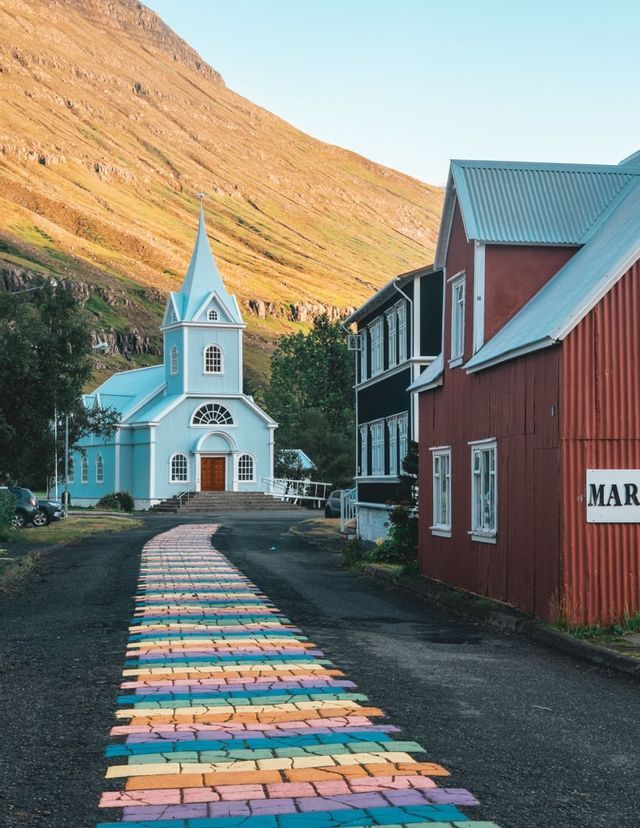 Ein regenbogenfarbener, bemalter Weg auf einer Asphaltstraße führt zu einer hellblauen Kirche am Fuße eines sonnenbeschienenen Berges.