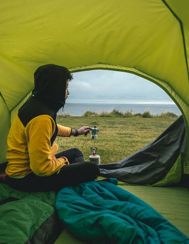 Una persona con una sudadera amarilla y negra prepara café en una cafetera moka, sentada dentro de una tienda de campaña con vistas al mar.