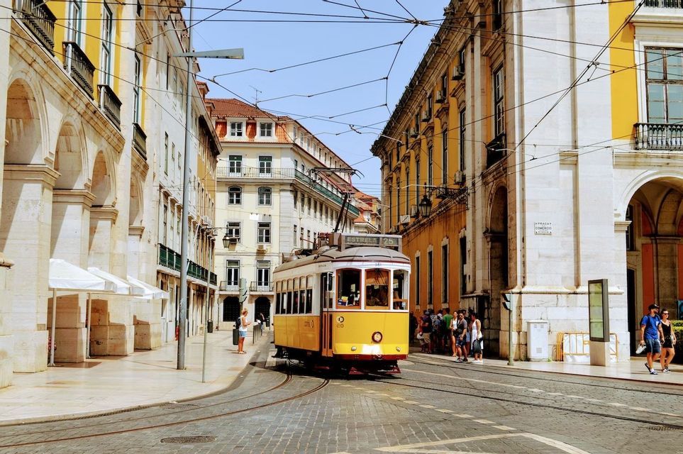 Un tram giallo percorre una strada lastricata che curva tra alti edifici classici europei sotto un cielo sereno.