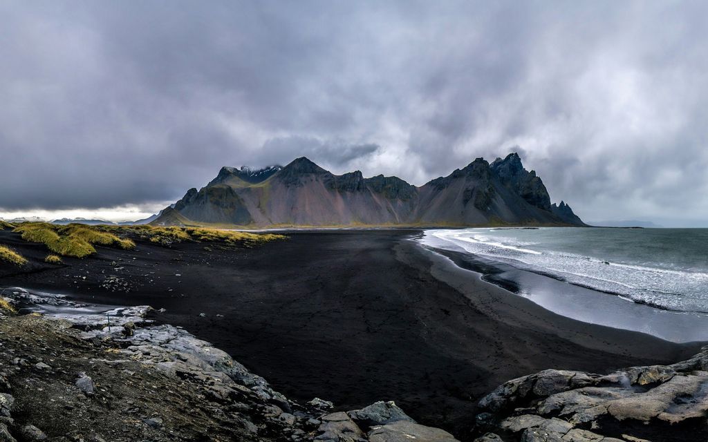 A panoramic view of a black sand beach with ocean waves meeting the shore in front of a rugged mountain range under a cloudy sky.