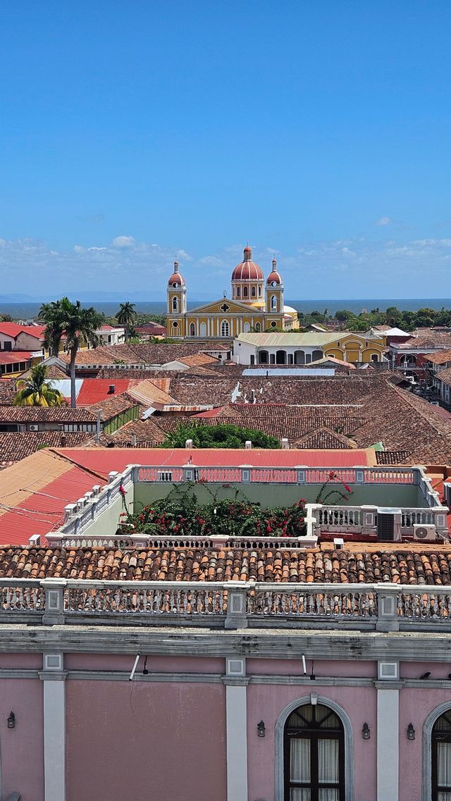Eine gelbe Kathedrale mit roten Kuppeln erhebt sich über die terrakottafarbenen Dächer einer Stadt, von einem hohen Aussichtspunkt aus gesehen, mit Wasser in der Ferne.