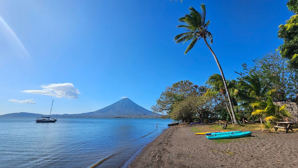 Ein Segelboot auf einem See vor einem Vulkan, vom dunklen Sandstrand aus gesehen, mit Kajaks und Palmen unter einem klaren blauen Himmel.
