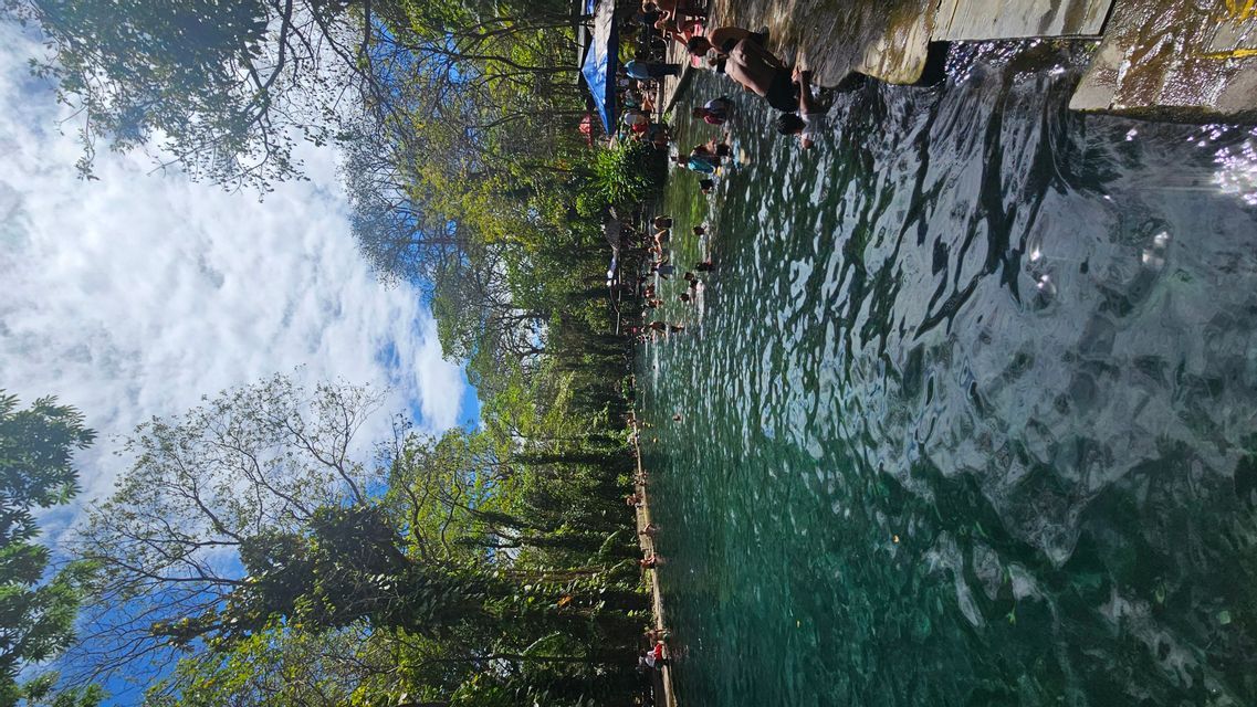 Eine WeRoad-Gruppenreise schwimmt und entspannt in einem Naturpool, umgeben von dichten grünen Bäumen, unter einem teilweise bewölkten Himmel.