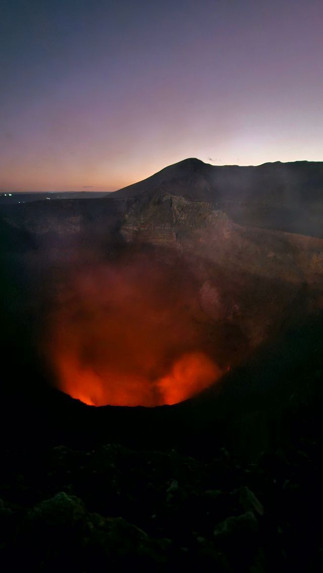 Ein tiefer Vulkankrater leuchtet in der Dämmerung mit rotglühender Lava und Rauch vor einer dunklen Bergsilhouette und einem violetten Himmel.