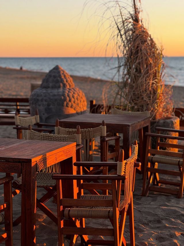 Tavoli e sedie di legno vuoti su una spiaggia sabbiosa al tramonto, con una grande testa di Buddha in pietra e l'oceano sullo sfondo.