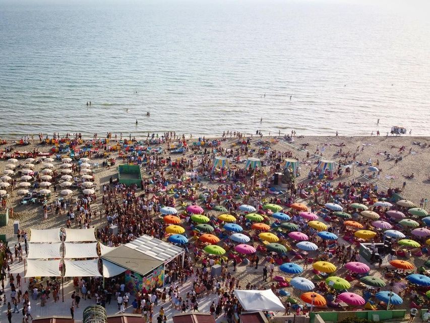 Una vista aerea di una festa in spiaggia affollata con persone sotto ombrelloni colorati e che nuotano nel mare calmo.