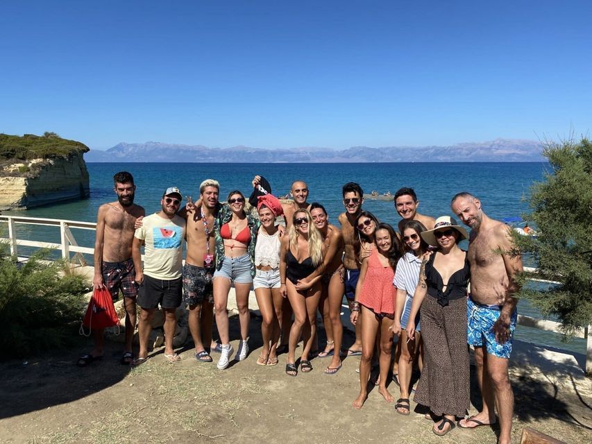 Eine große WeRoad Gruppenreise posiert für ein Foto an einem sonnigen Strand, mit einer felsigen Klippe und dem blauen Meer im Hintergrund.