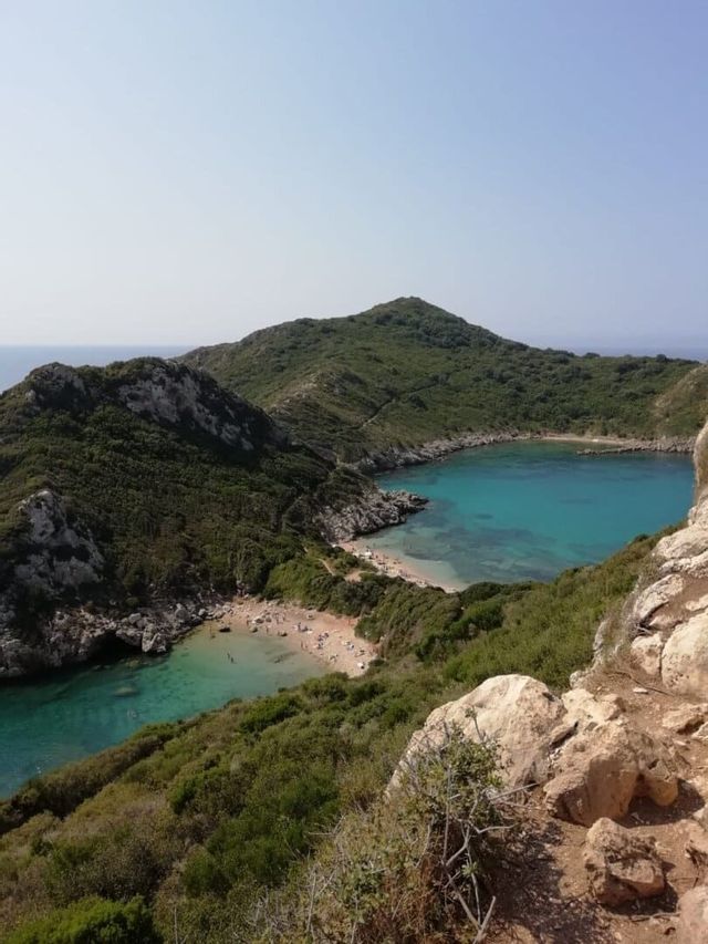 Vista aérea de una cala doble con aguas turquesas y playas de arena, enclavada entre exuberantes colinas verdes bajo un cielo despejado.