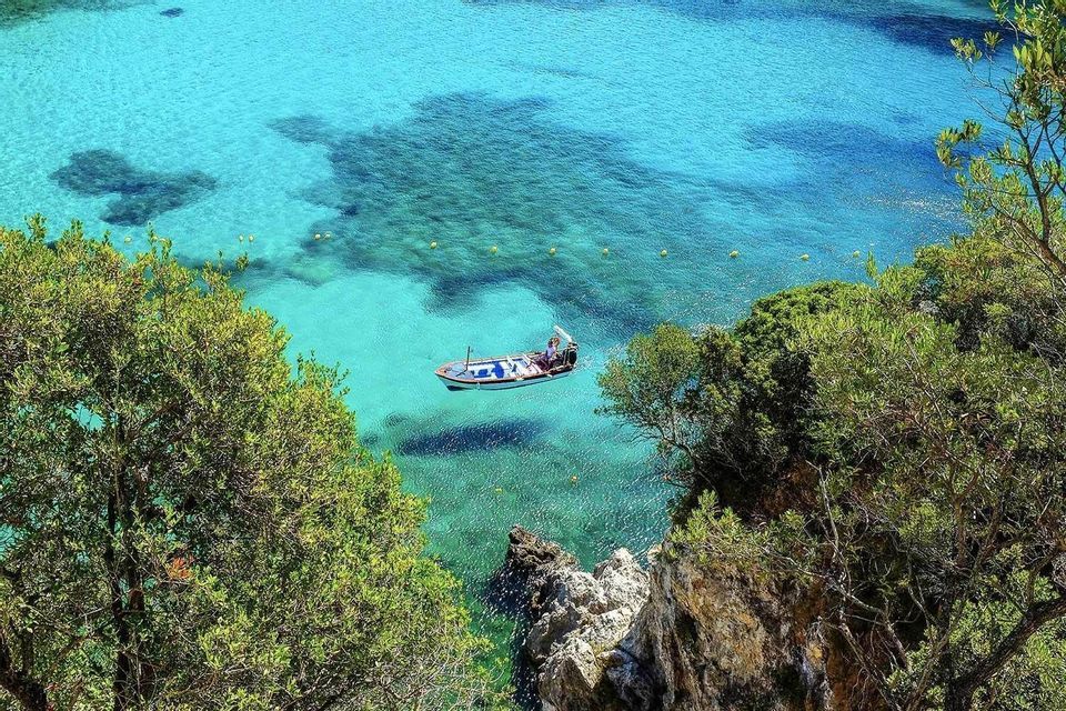 Vue en plongée d'un petit bateau flottant sur une eau turquoise cristalline, aperçu entre des arbres verts le long d'une côte rocheuse.