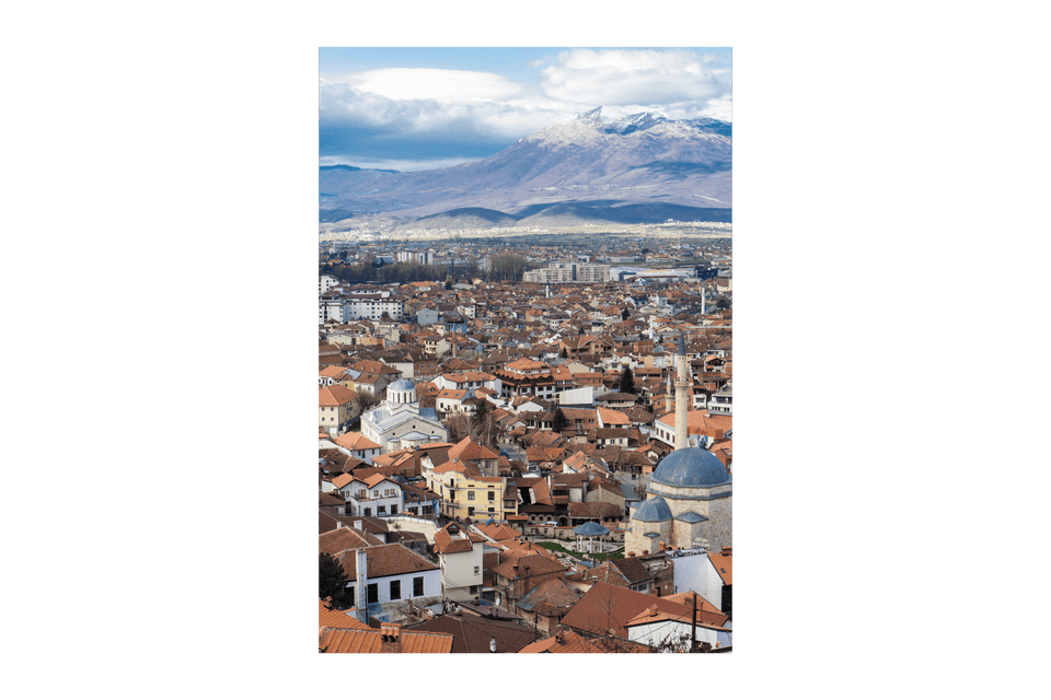 Veduta aerea di una città con tetti di tegole rosse, con montagne innevate che si ergono sullo sfondo sotto un cielo parzialmente nuvoloso.