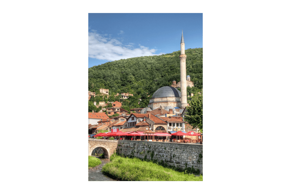 Una moschea con cupola e minareto domina una cittadina con edifici dai tetti rossi. In primo piano, un ponte di pietra attraversa un fiume.