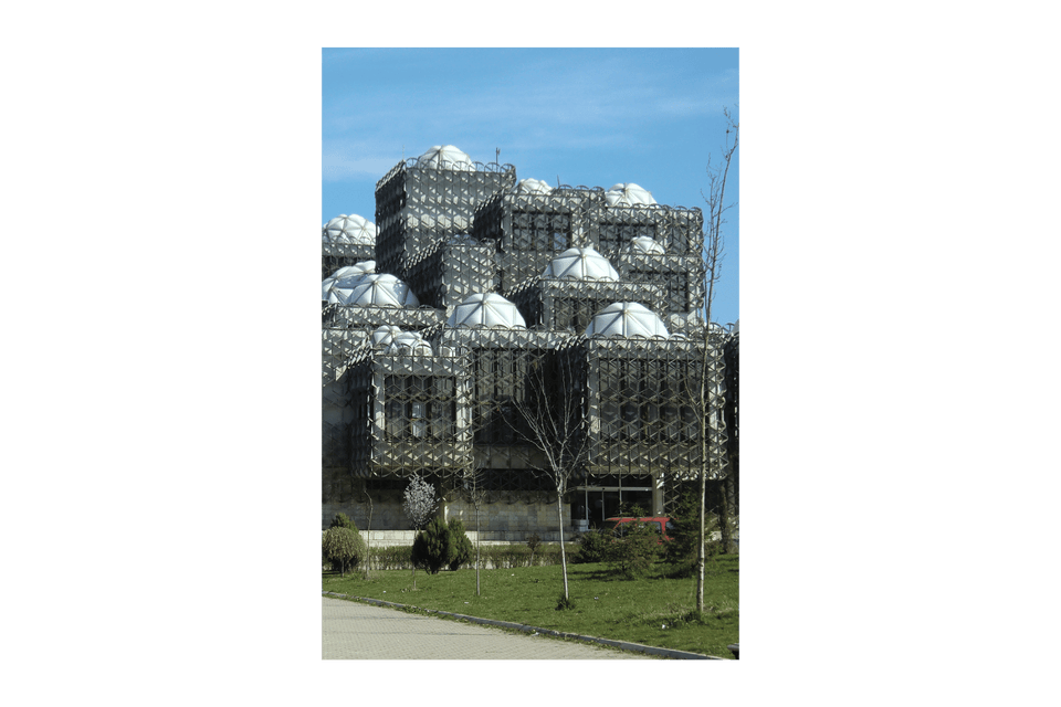 Un edificio brutalista con cubi impilati, una facciata a traliccio metallico e cupole bianche, situato su un prato verde sotto un cielo sereno.