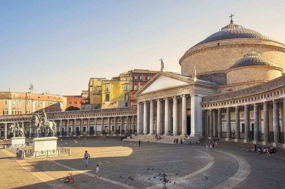 Une place publique ensoleillée, ornée d'un grand bâtiment à dôme et d'une colonnade classique, avec des passants et des statues dispersés.