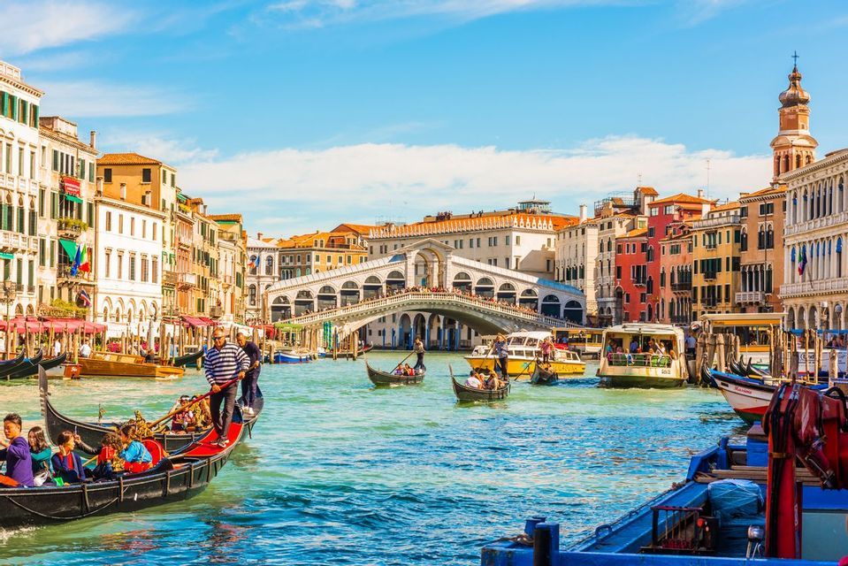 Des gondoles et d'autres bateaux naviguent sur un canal animé, avec un grand pont de pierre voûté et des bâtiments historiques colorés sous un ciel bleu.