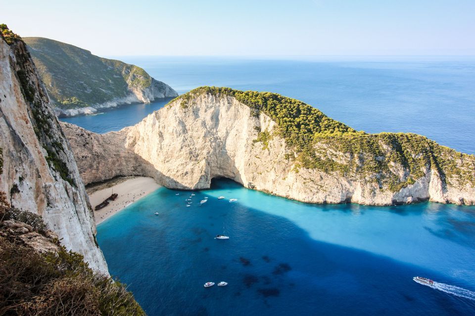 Vista aérea de una cala apartada con un naufragio en una playa de arena blanca, rodeada de acantilados y agua turquesa con barcos.