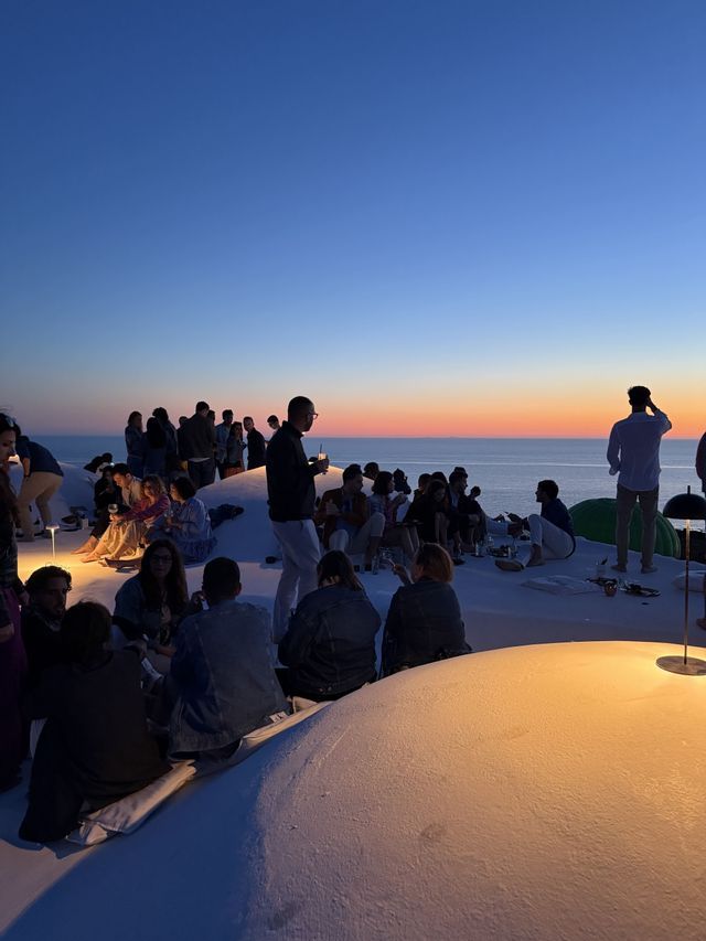 Un gruppo WeRoad socializza su un tetto bianco, ammirando il tramonto sul mare.