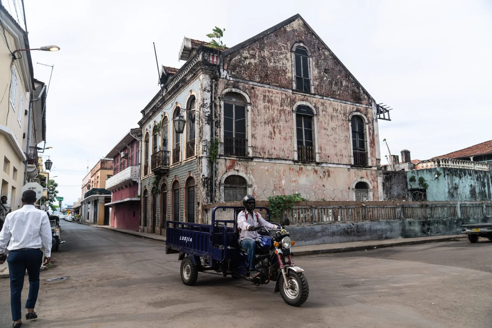 Un uomo percorre una strada a bordo di una moto a tre ruote blu, passando accanto a vecchi edifici in stile coloniale segnati dal tempo, sotto un cielo coperto.