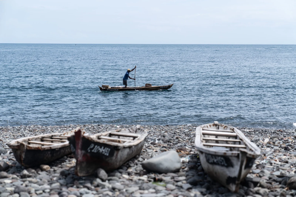 Un uomo su una barca di legno rema in mare, mentre altre tre barche sono parcheggiate su una spiaggia di ciottoli in primo piano.