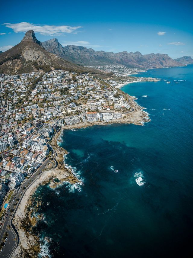Une vue aérienne d'une ville sur un littoral rocheux, avec une chaîne de montagnes en arrière-plan sous un ciel bleu.