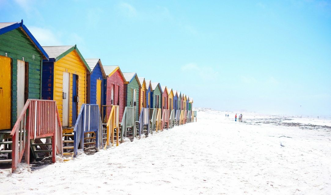 Une rangée de cabanes de plage en bois colorées se dressant sur une plage de sable blanc sous un ciel bleu clair.