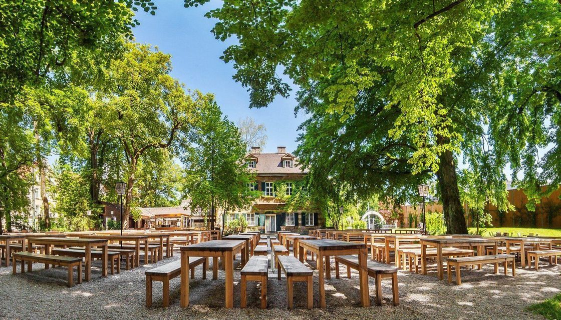 File di tavoli e panche di legno vuoti si trovano su un patio ghiaioso in una birreria all'aperto, ombreggiata da grandi alberi frondosi.