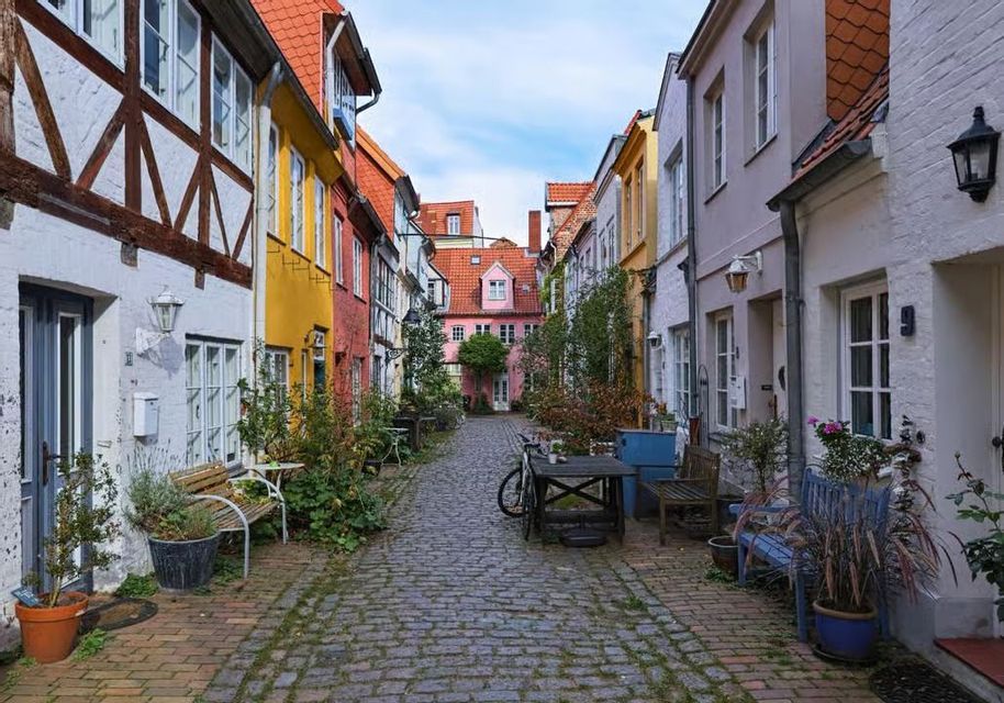 Un vicolo stretto di ciottoli fiancheggiato da case tradizionali e colorate, piante in vaso e mobili da esterno sotto un cielo parzialmente nuvoloso.