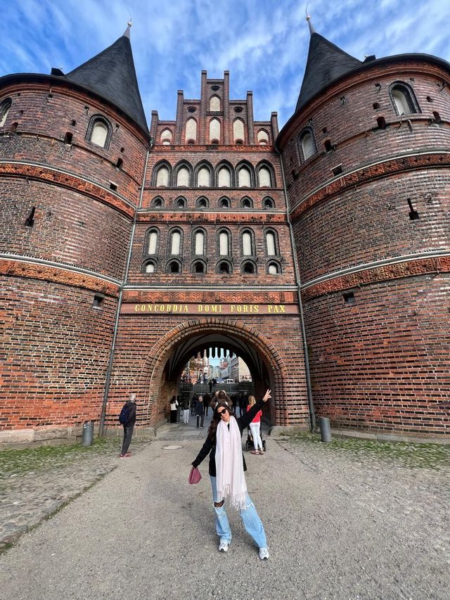 Una donna in jeans strappati e una sciarpa bianca posa davanti a un casello storico di mattoni rossi con torri gemelle.