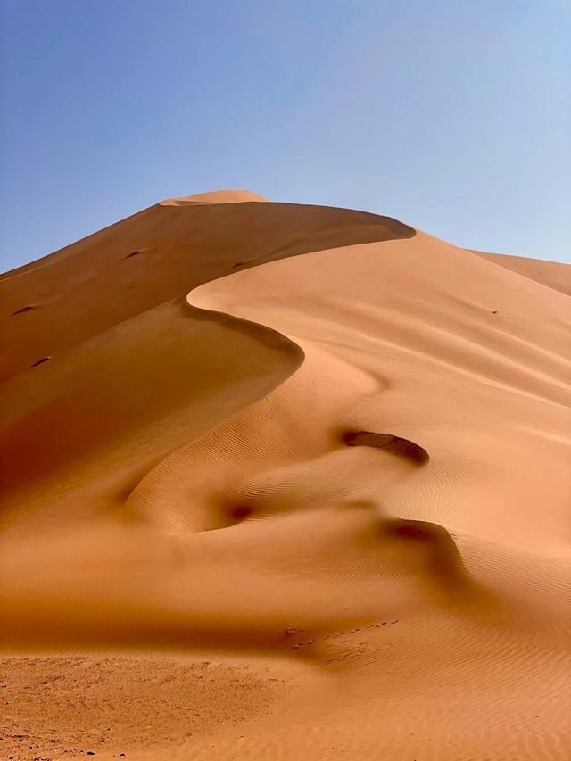 La netta curva a S della cresta di una grande duna di sabbia arancione crea giochi di luce e ombra sotto un cielo azzurro e limpido.