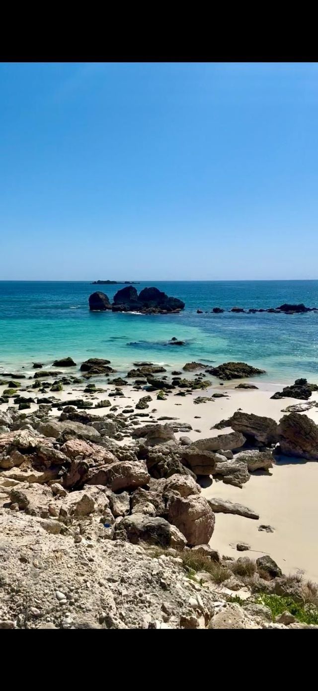 Una costa rocciosa degrada in una spiaggia di sabbia bianca, lambita da acque turchesi e blu, sotto un cielo sereno.