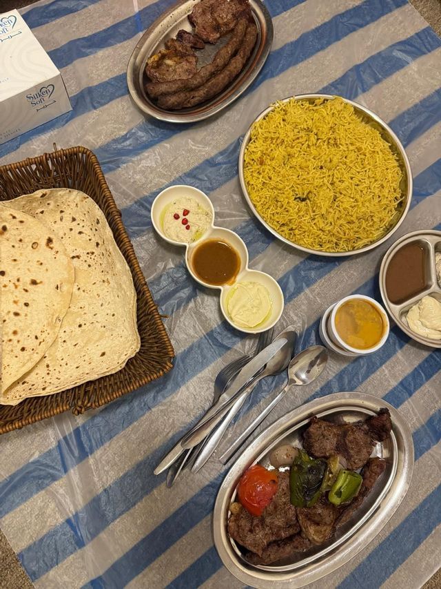Un pasto tradizionale a base di riso giallo, carne alla griglia, pane azzimo e salse è servito su una tovaglia a righe.