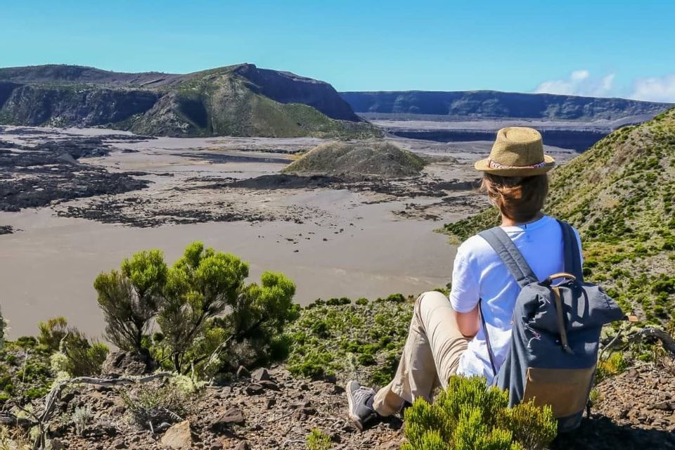 Una persona con uno zaino e un cappello è seduta su un'altura verde che si affaccia su una vasta caldera vulcanica in una giornata di sole.