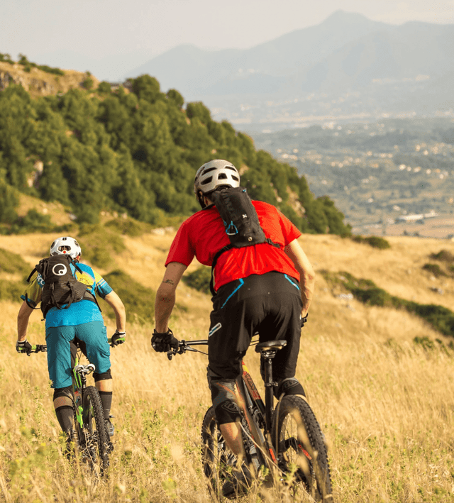 Due ciclisti percorrono in mountain bike un sentiero erboso in discesa, con una valle e montagne visibili in lontananza.