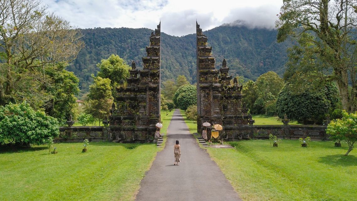 A woman seen from behind walks on a path towards an ornate split stone gate, with lush green trees and a mountain in the background.