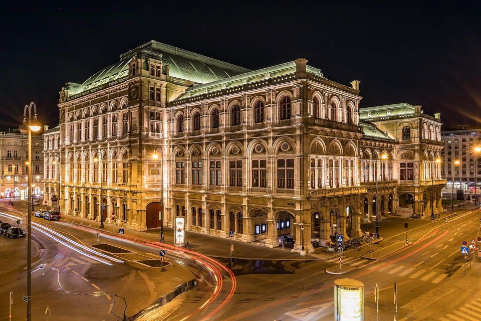 Un bâtiment historique orné, illuminé la nuit au coin d'une rue de ville, avec des traînées lumineuses dues au trafic en pose longue.