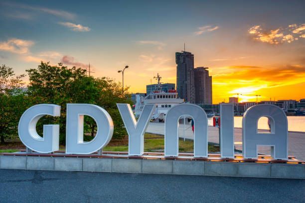 Una grande insegna bianca con la scritta 'GDYNIA' su una piattaforma in riva all'acqua, con lo skyline della città e un tramonto colorato sullo sfondo.