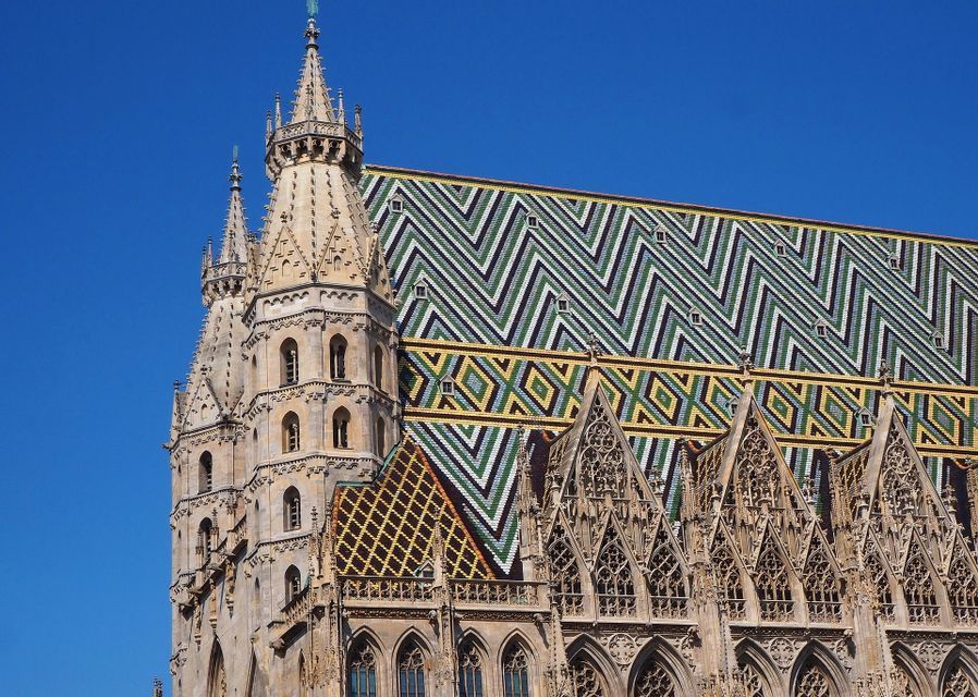 Gros plan sur la flèche de pierre ornée et le toit de tuiles colorées aux motifs géométriques d'une cathédrale, sous un ciel bleu clair.