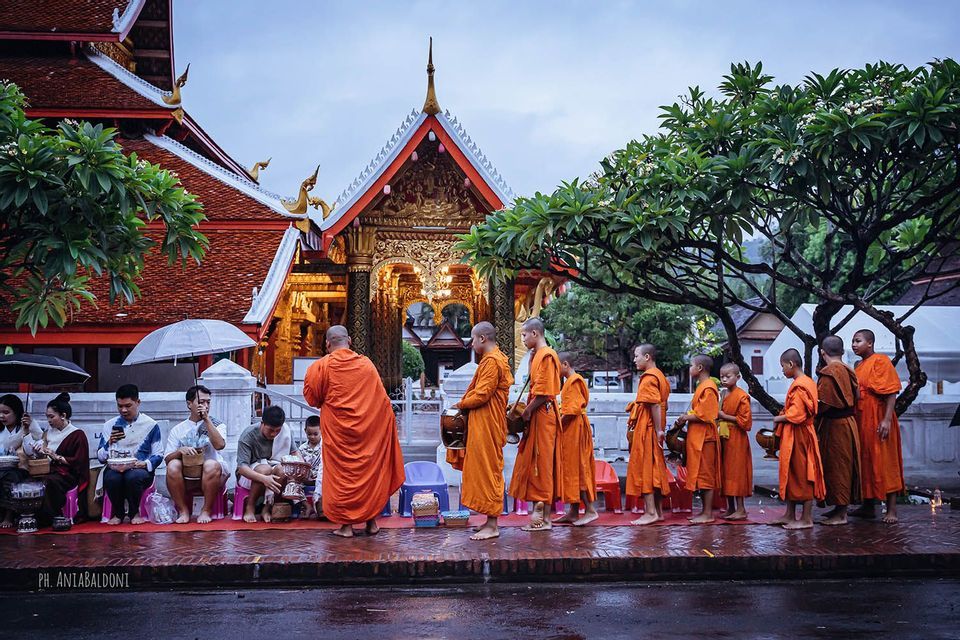 Monaci buddisti in veste arancione si allineano per ricevere elemosine da persone sedute davanti a un tempio ornato.