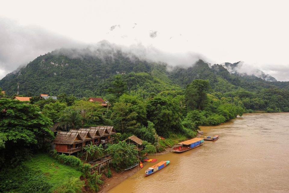 Palafitte in legno e barche longtail riposano sulla riva di un fiume fangoso ai piedi di una montagna coperta di nuvole.