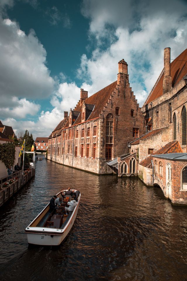 Un bateau de tourisme transportant des passagers navigue sur un canal, longeant des bâtiments historiques en briques rouges aux toits à pignon, sous un ciel nuageux.