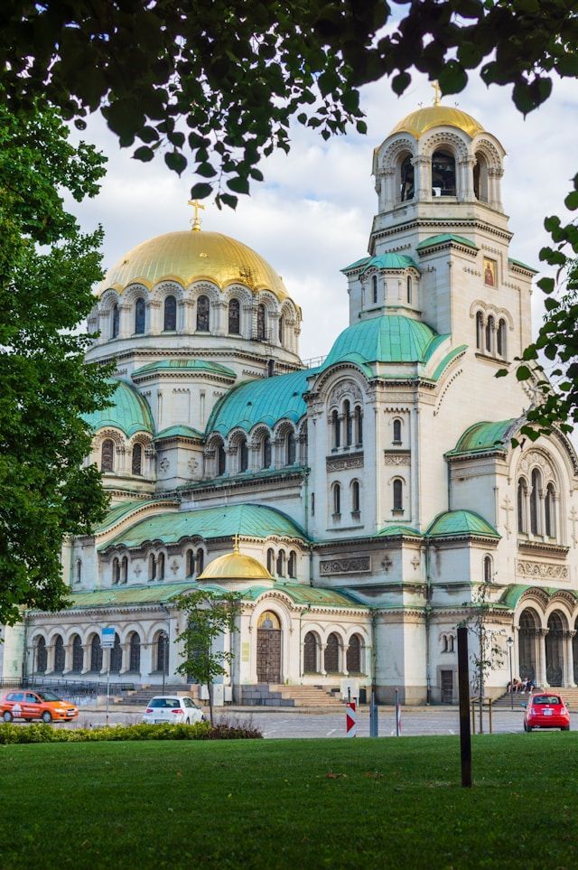 Eine prächtige weiße Kathedrale mit großen goldenen und kleineren türkisfarbenen Kuppeln, gesehen hinter den Blättern grüner Bäume.