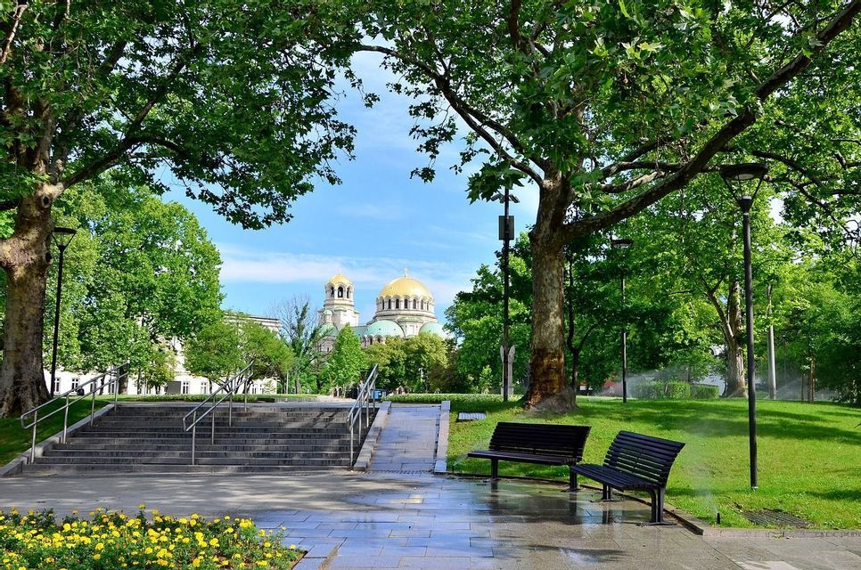 Ein Blick aus einem üppigen Stadtpark mit Steinwegen und Bänken, der eine ferne Kathedrale mit großen goldenen Kuppeln unter blauem Himmel umrahmt.