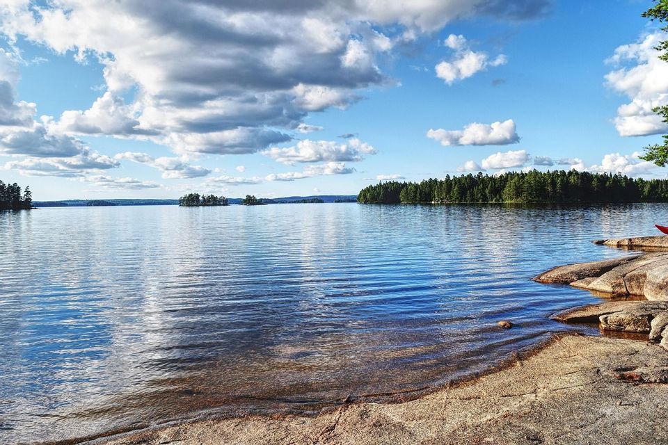 Ein breiter, ruhiger See mit felsigem Ufer unter einem teilweise bewölkten blauen Himmel, mit kieferbewachsenen Inseln in der Ferne.