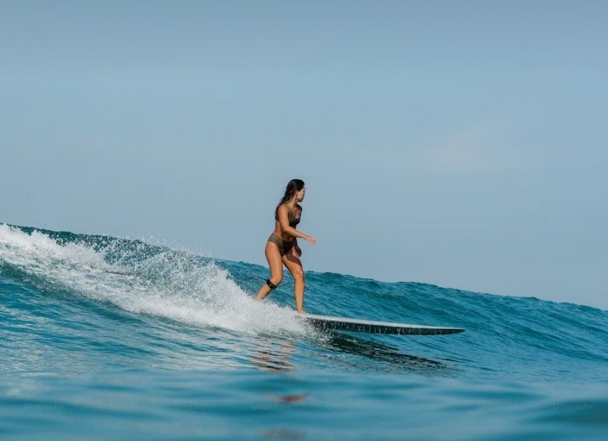 Eine Frau im Bikini mit Kniebandage surft auf einem Longboard auf einer blauen Ozeanwelle unter klarem Himmel.