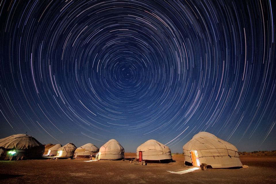 Una fila de yurtas iluminadas se asienta en el suelo por la noche bajo un cielo con rastros circulares de estrellas de larga exposición.