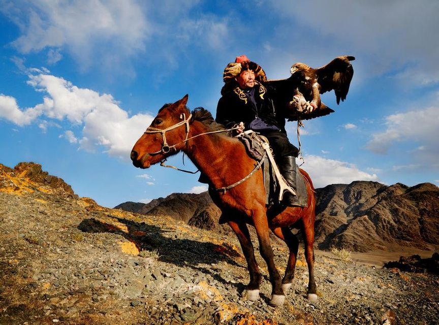 Una persona con vestimenta tradicional a caballo sostiene una gran águila en su brazo en una ladera rocosa bajo un cielo parcialmente nublado.